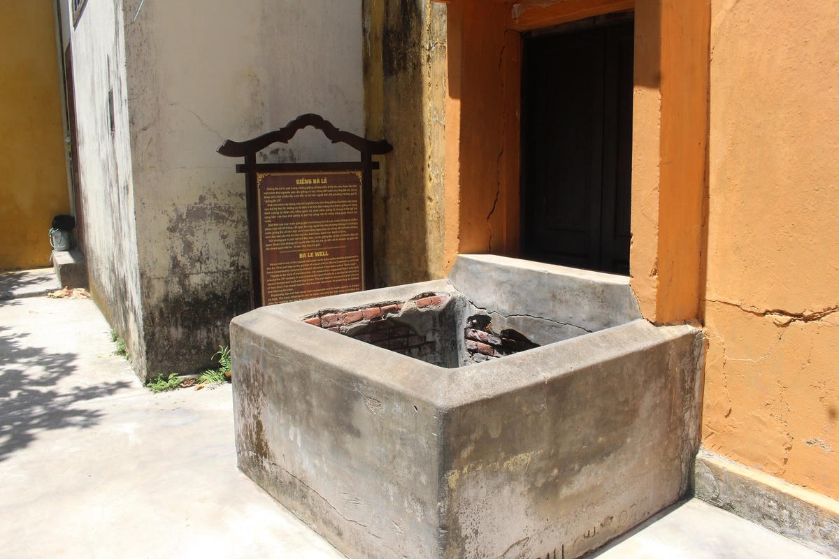 Bá Lễ well seen from above, its square concrete rim with one corner worn through to older brick beneath, a bilingual wooden sign reading GIẾNG BÁ LỄ and BA LE WELL standing beside it against an orange-painted wall, no one in view.