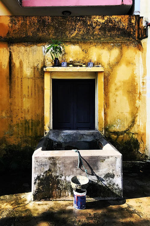 The square brick rim of Bá Lễ well against a mustard-yellow wall stained with moss, a small shrine shelf fixed above it holding a potted plant, two small jars and an incense bowl, a paint bucket standing beside the well.