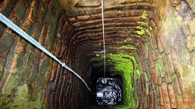 Looking straight down the shaft of Bá Lễ well, its wall built from rings of moss-covered red brick, a rope for lowering a bucket disappearing into dark water far below.