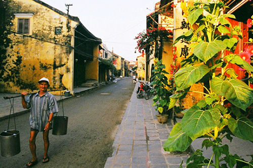 A man in a conical hat carries two water buckets on a shoulder pole down a stone-paved street in Hội An's old quarter, tile-roofed houses and pots of red bougainvillea along both sides.