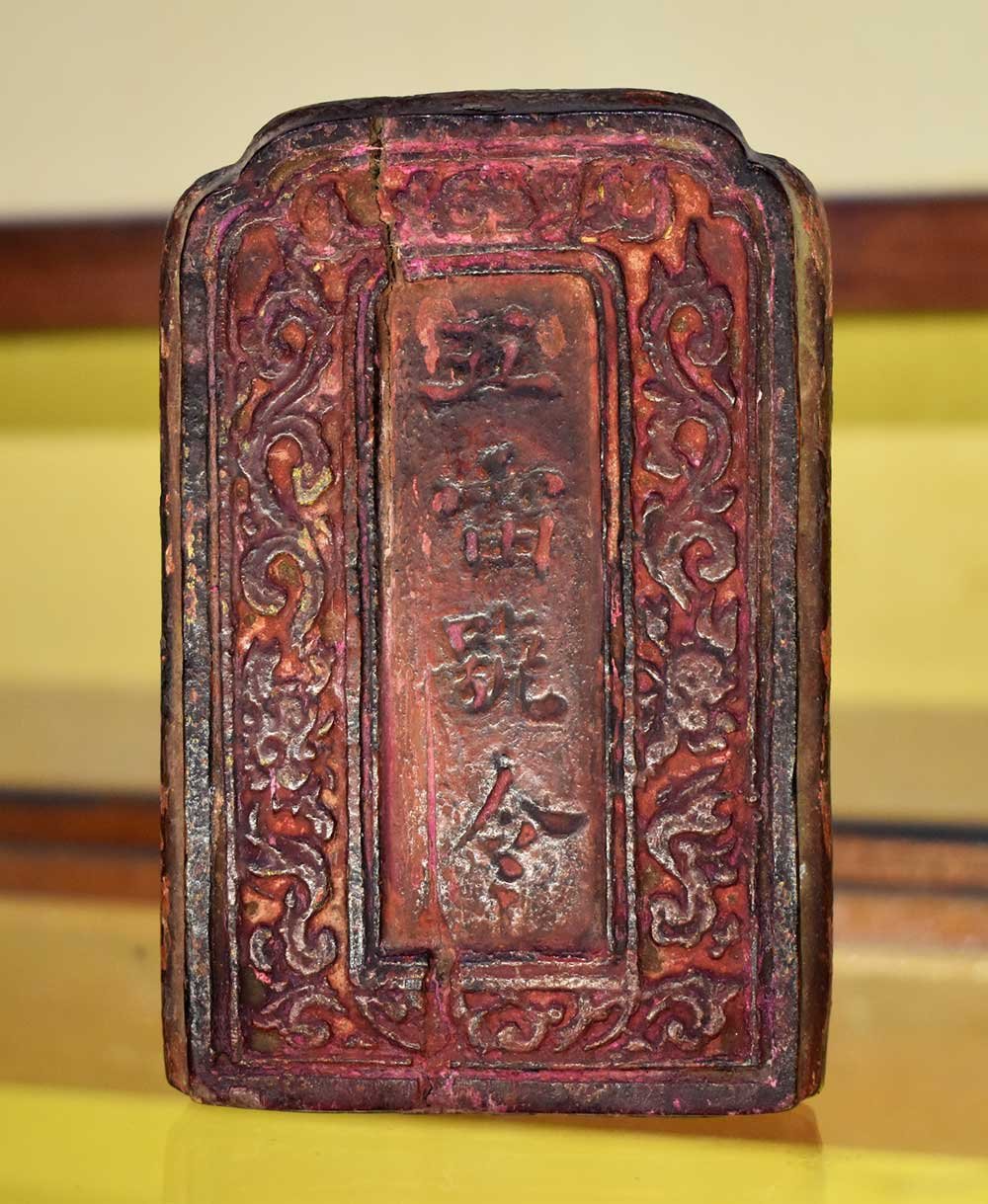 Mặt trước tấm lệnh phù: khối gỗ sơn đỏ đã phai màu, đỉnh cong hình vòng cung, viền ngoài chạm hoa văn dây leo nổi, chính giữa là ô chữ nhật khắc bốn chữ Hán theo cột dọc, một vết nứt chạy dọc hiện vật.