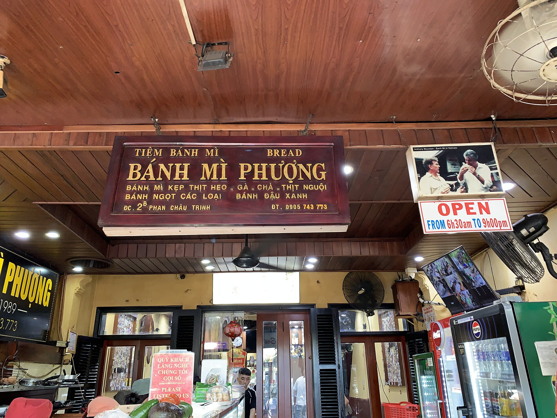 Bảng hiệu gỗ 'Tiệm bánh mì Bánh mì Phượng' treo trên cửa tiệm; bên phải bảng có khung ảnh gắn đèn, trong ảnh hai người đàn ông đang ăn bánh mì, dòng chữ tiếng Anh phía trên ảnh nhắc tên Anthony Bourdain.