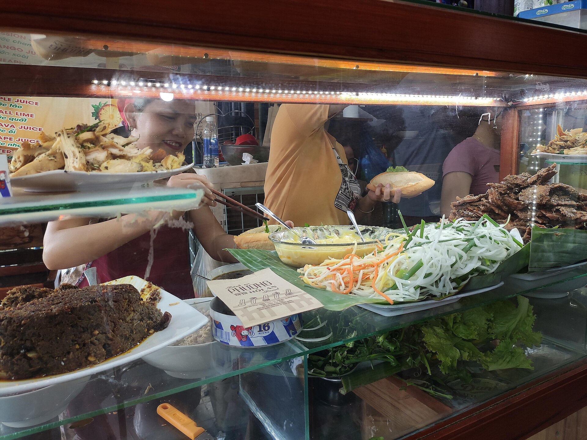 Nhìn qua tấm kính một tủ bánh mì: người phụ nữ sau quầy dùng đũa gắp thức ăn, giữa quầy có tô trứng, hành và cà rốt thái sợi xếp trên lá chuối, một ổ bánh nằm cạnh.