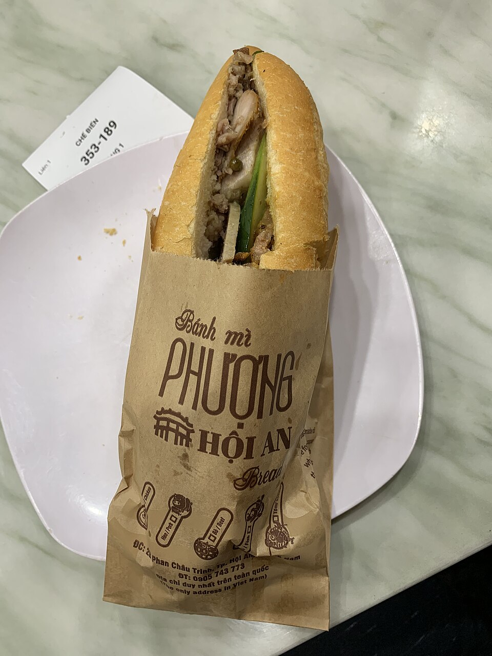 Một ổ bánh mì cắt dọc trong túi giấy nâu in chữ 'Bánh mì Phượng Hội An' đặt trên đĩa; ruột ổ nhồi thịt, dưa leo và chả, đầu bánh nhọn.