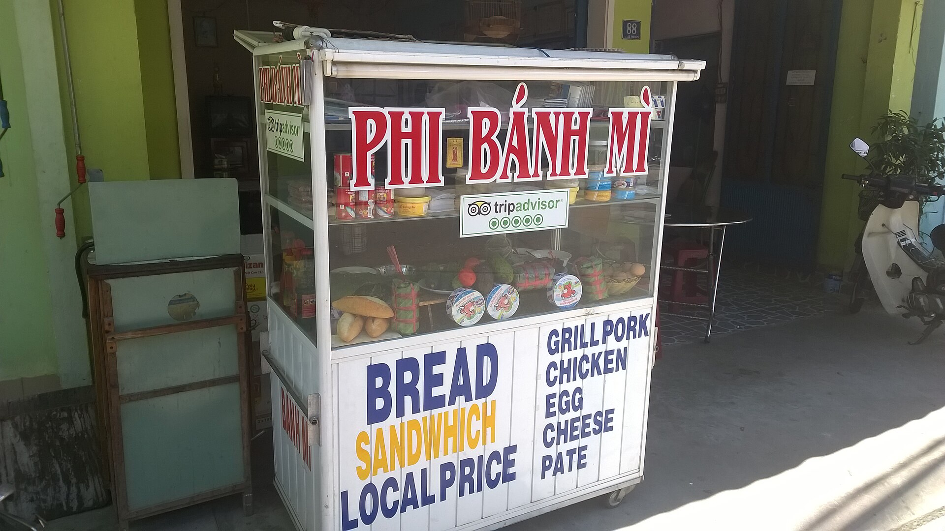 Một tủ bánh mì kính đặt bên đường, bảng trắng chữ đỏ 'PHI BÁNH MÌ', mặt trước ghi chữ tiếng Anh về bánh mì, giá bán và danh sách nhân; trong tủ có vài ổ bánh, hộp phô mai, lon đồ hộp và rau.