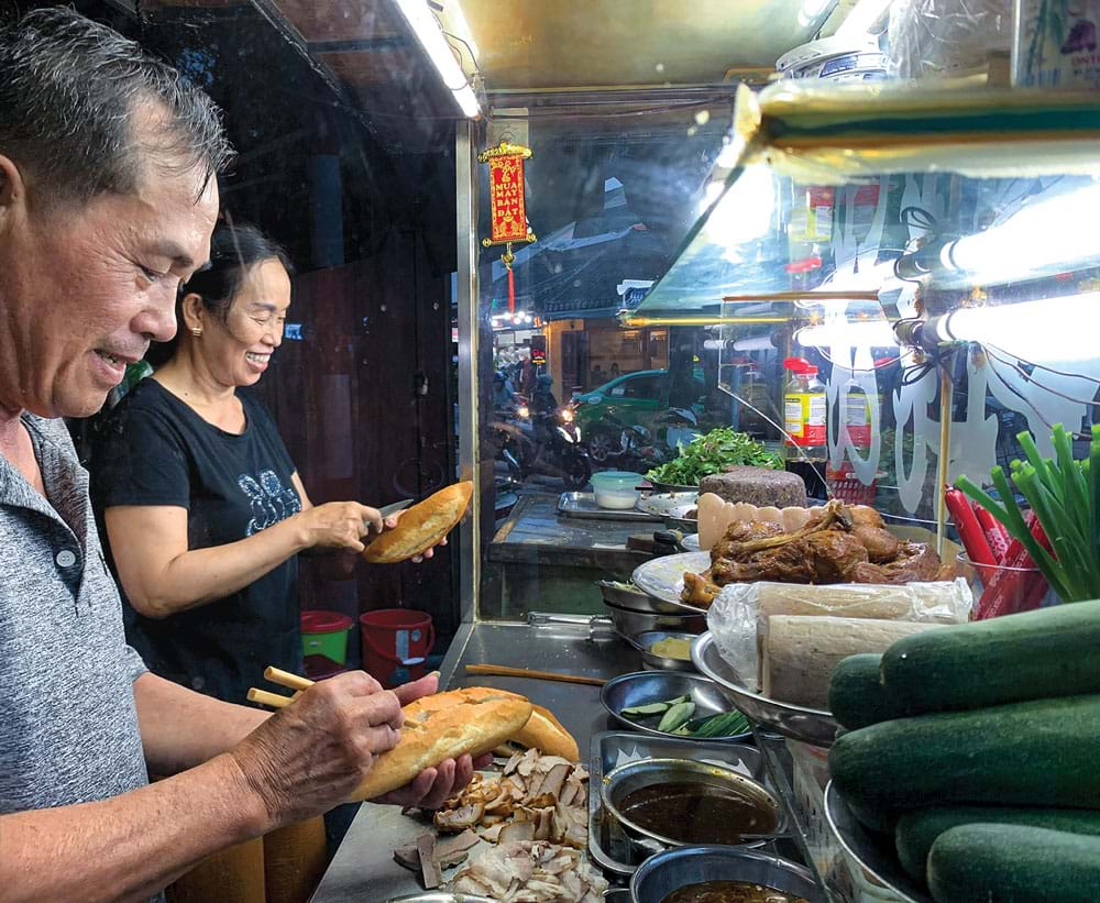 Hai người đứng sau một tủ bánh mì kính ở ngã tư về đêm: người đàn ông áo xám rạch một ổ bánh, người phụ nữ áo đen phía sau cầm một ổ khác; trên quầy có thịt xíu thái, thịt quay, chả, dưa leo, hành lá và ớt.