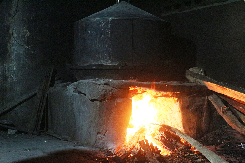 Close on the mouth of a cao lầu noodle furnace at night: firewood burns bright orange under a cone-topped tin steaming pot, split logs stacked to one side.