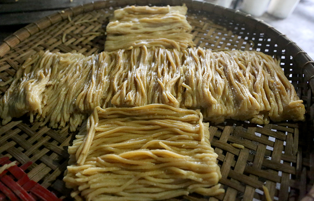 Pale yellow skeins of cao lầu noodles arranged in a cross on a woven bamboo tray, a scatter of sliced red chilli visible at one edge.