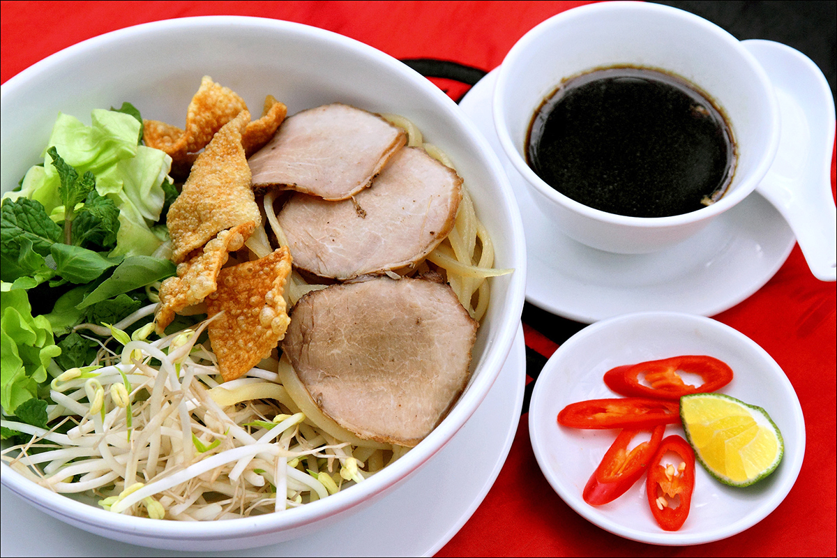 A bowl of cao lầu seen from above: noodles under lettuce, mint, bean sprouts, crisp fried crackers and three slices of char siu pork, beside a small cup of dark sauce and a dish of chilli and lime.