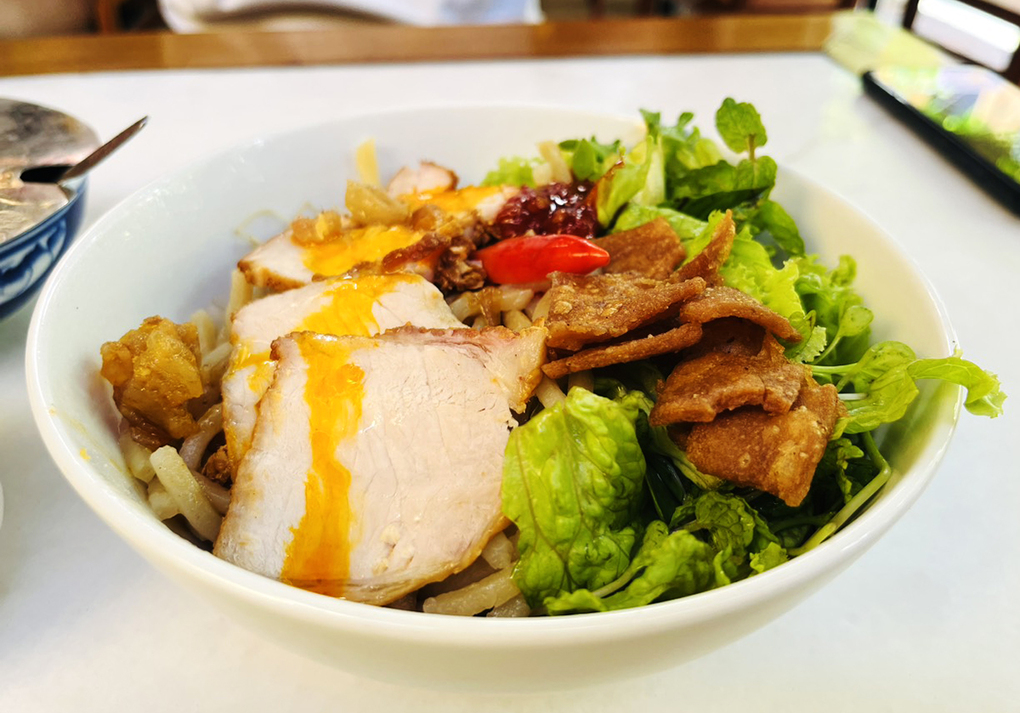 A bowl of cao lầu seen from the side: three slices of char siu drizzled with orange sauce next to lettuce, mint, bean sprouts and crisp fried pork skin, one red chilli resting in the middle.