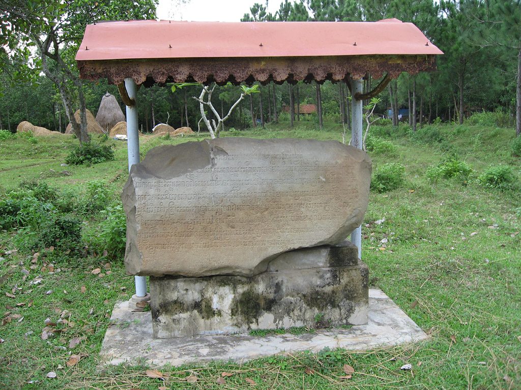 A large inscribed boulder, cracked through the middle, standing under an open-sided roof at Chiên Đàn, rice fields and straw stacks behind it.