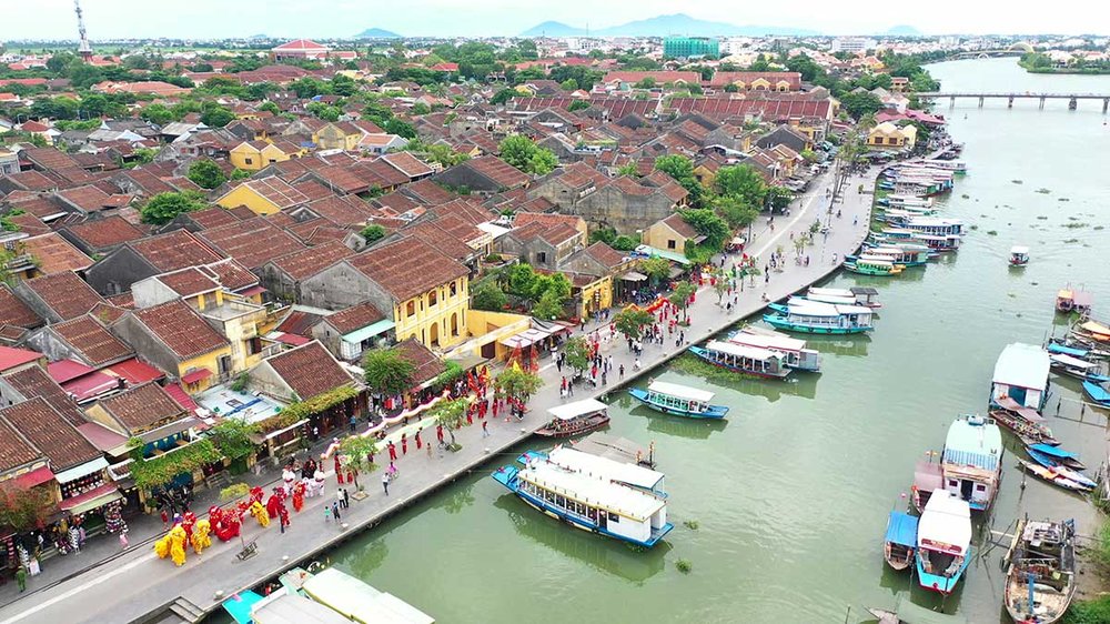 An aerial view of Hội An's riverside road during the Quan Công festival: a procession in red robes with a lion and dragon dance troupe moves along the road beside the Hoài river, tourist boats moored below and the old town's tiled roofs stretching back from the water.