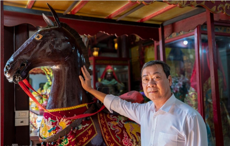 A man in his sixties rests one hand on the neck of a life-sized dark wooden horse statue draped in an embroidered red and gold saddle cloth, standing inside a red-lacquered shrine with a seated painted figure visible behind glass.