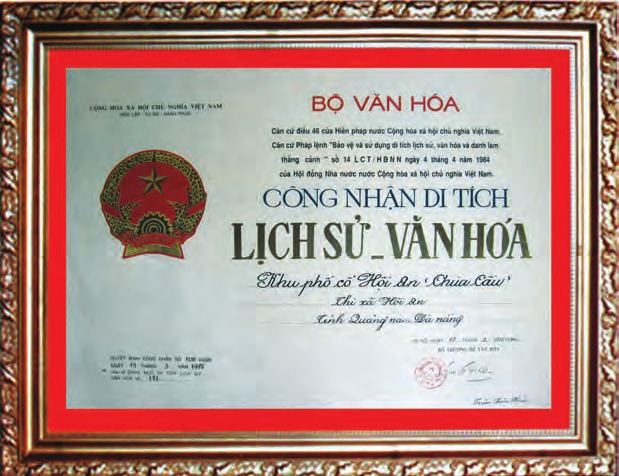A certificate in a gilt wooden frame with a red mount: the state emblem of Vietnam at the left, BỘ VĂN HÓA in red above, then CÔNG NHẬN DI TÍCH LỊCH SỬ - VĂN HÓA in heavy type, and below it in script “Khu phố cổ Hội An ‘Chùa Cầu’ · Thị xã Hội An · Tỉnh Quảng Nam - Đà Nẵng”. The line carrying the decree number at the lower left is too faint to read.