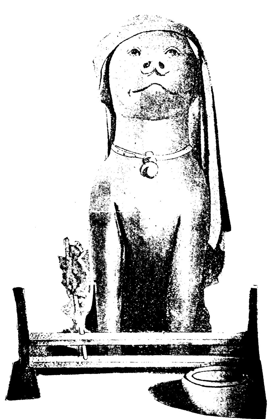 A black and white photograph of a seated dog figure on a wooden altar table, a cloth draped over its head and down both sides, a collar with a crescent-shaped pendant at its throat, a bowl and a sprig of leaves in front of it.
