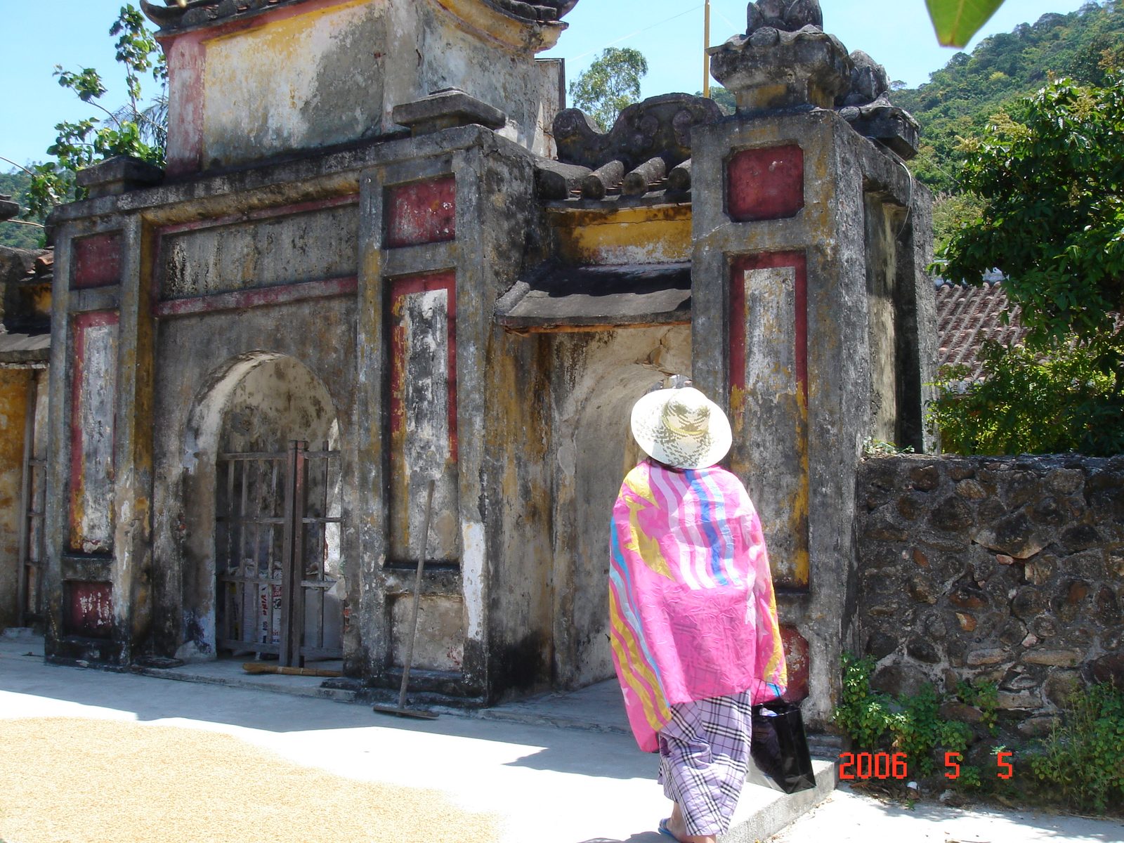 The weathered stone gate of an old pagoda on the island, red and ochre paint faded and stained, a visitor in a wide straw hat and a bright poncho walking toward the arched doorway, forested hills behind.