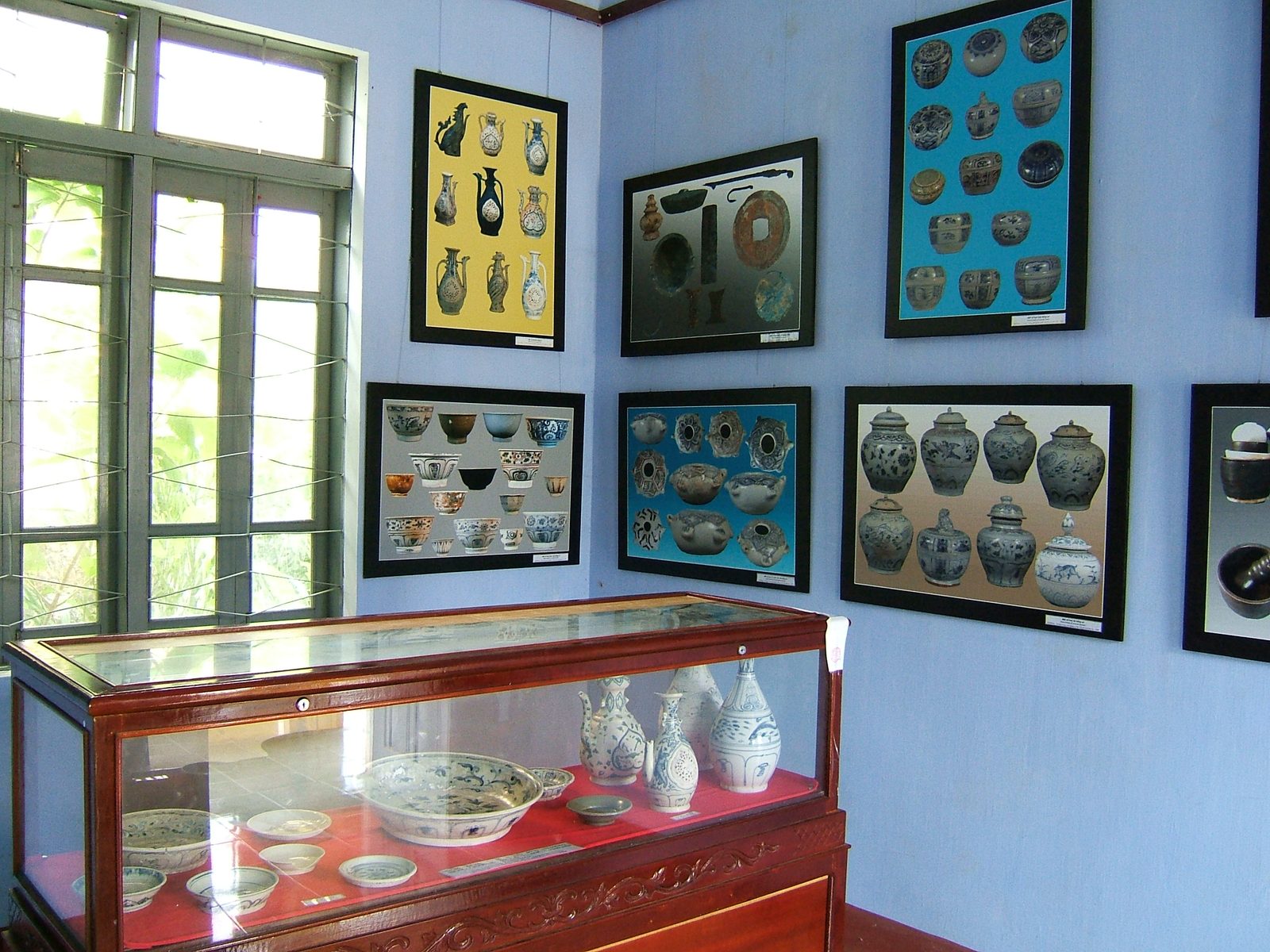 A museum display case holding blue-and-white ceramic bowls, dishes and a two-spouted ewer recovered from the shipwreck, with framed reference boards on the wall behind showing further examples of the ship's ceramic types.