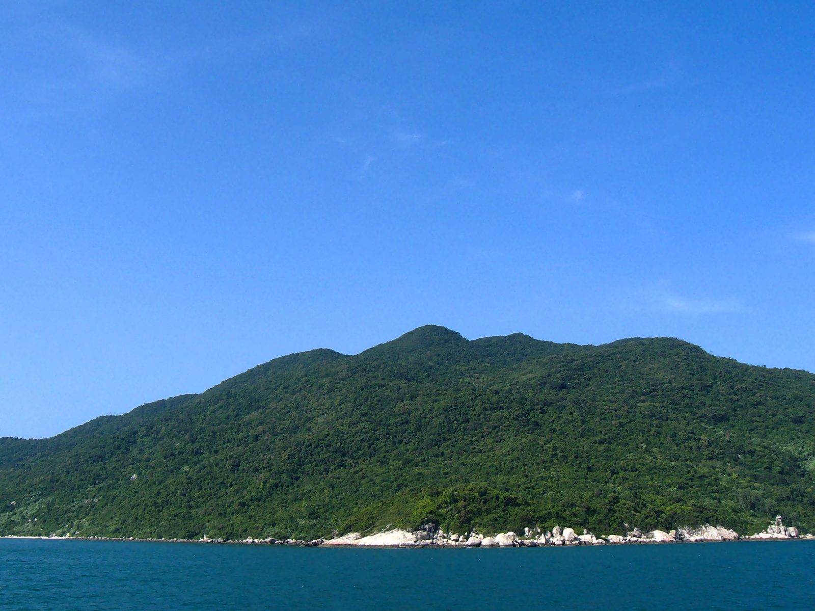 The forested ridge of Hòn Lao seen from the sea under a clear blue sky, pale granite boulders exposed along the shoreline, calm dark-blue water in the foreground.