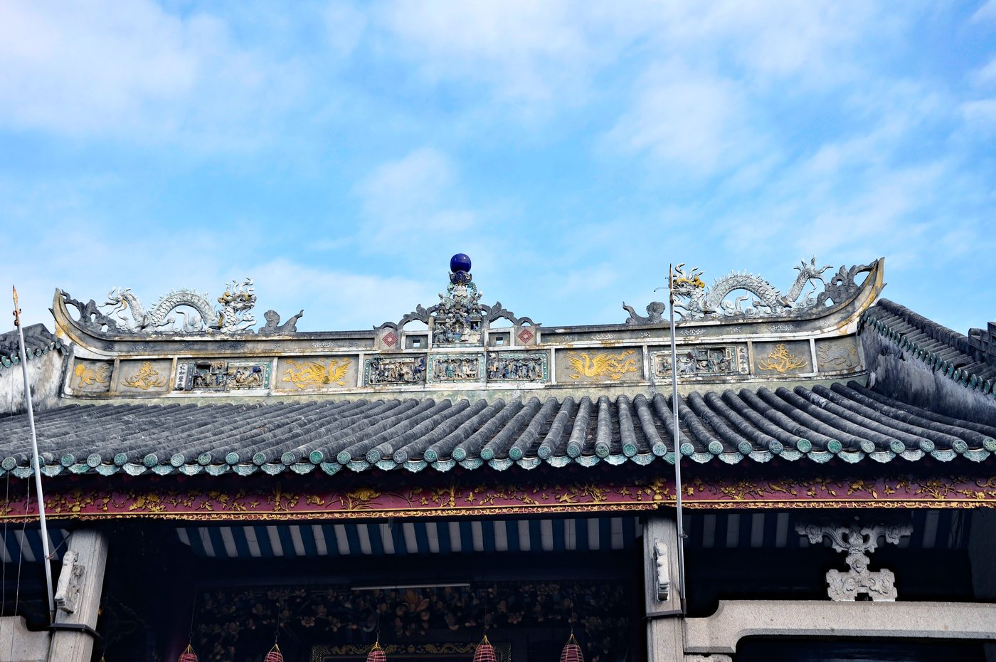 The roofline of the Cantonese hall in Hội An seen from below, a row of glazed ceramic figures along the ridge over tubular tiles, a dragon facing in at either end.
