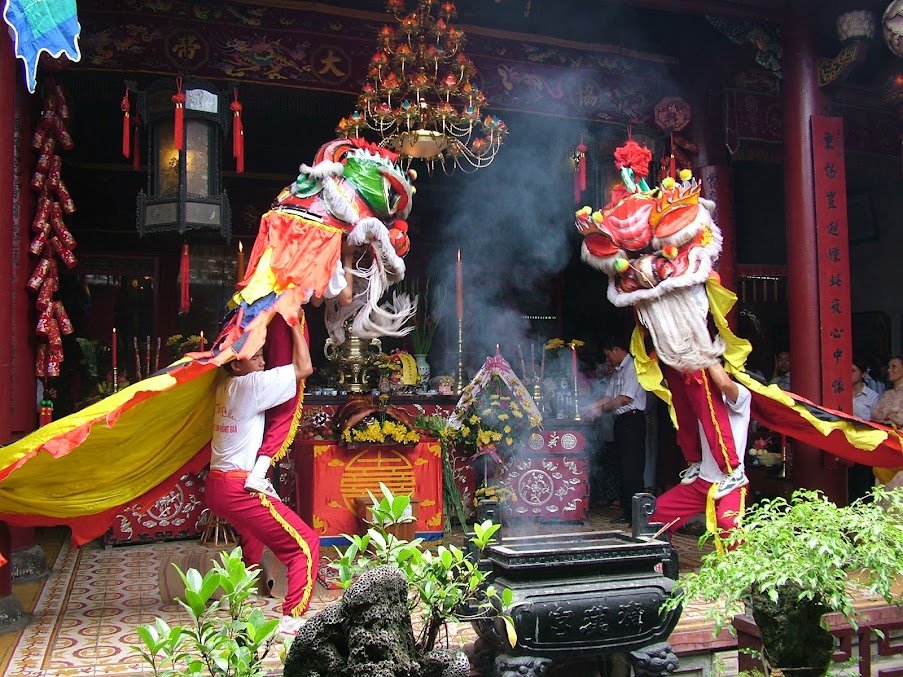 Hai đội múa lân nâng đầu lân cao trước bàn thờ khói hương nghi ngút, đèn chùm cổ và câu đối đỏ phía sau, chậu cây cảnh bày quanh sân.