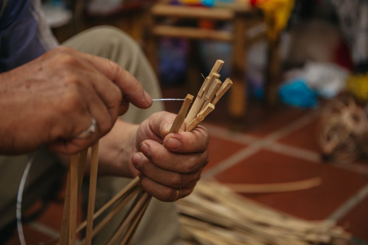 Close-up of a worker's two hands tying a bundle of sharpened bamboo strips with a thin cord, a pile of dried bamboo strips resting on the tiled floor below.