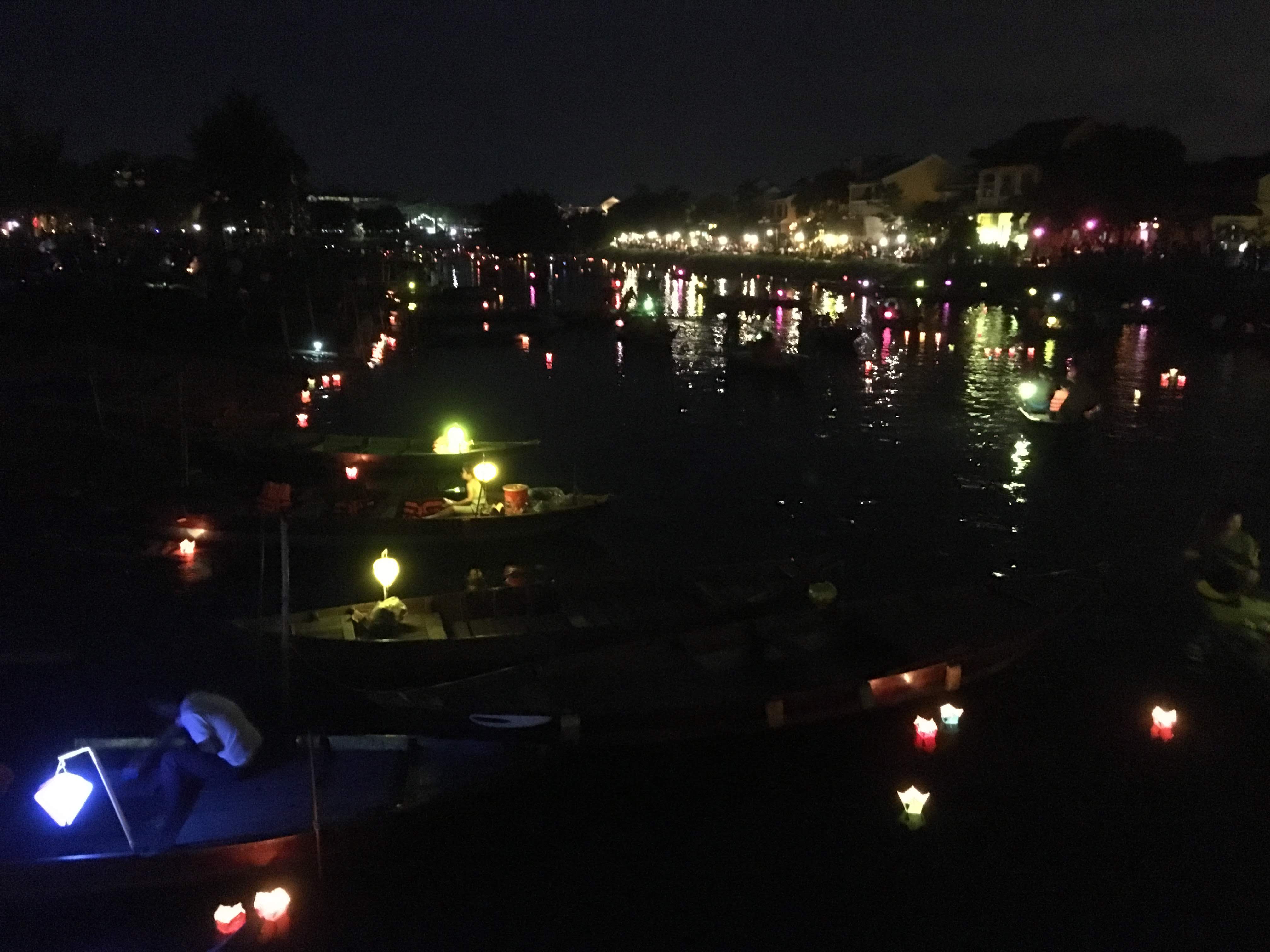 Small boats moored close together on the Hoài river at night, each hung with one or two small lit lanterns in yellow, orange and blue, the old quarter's lights reflected on the water beyond.