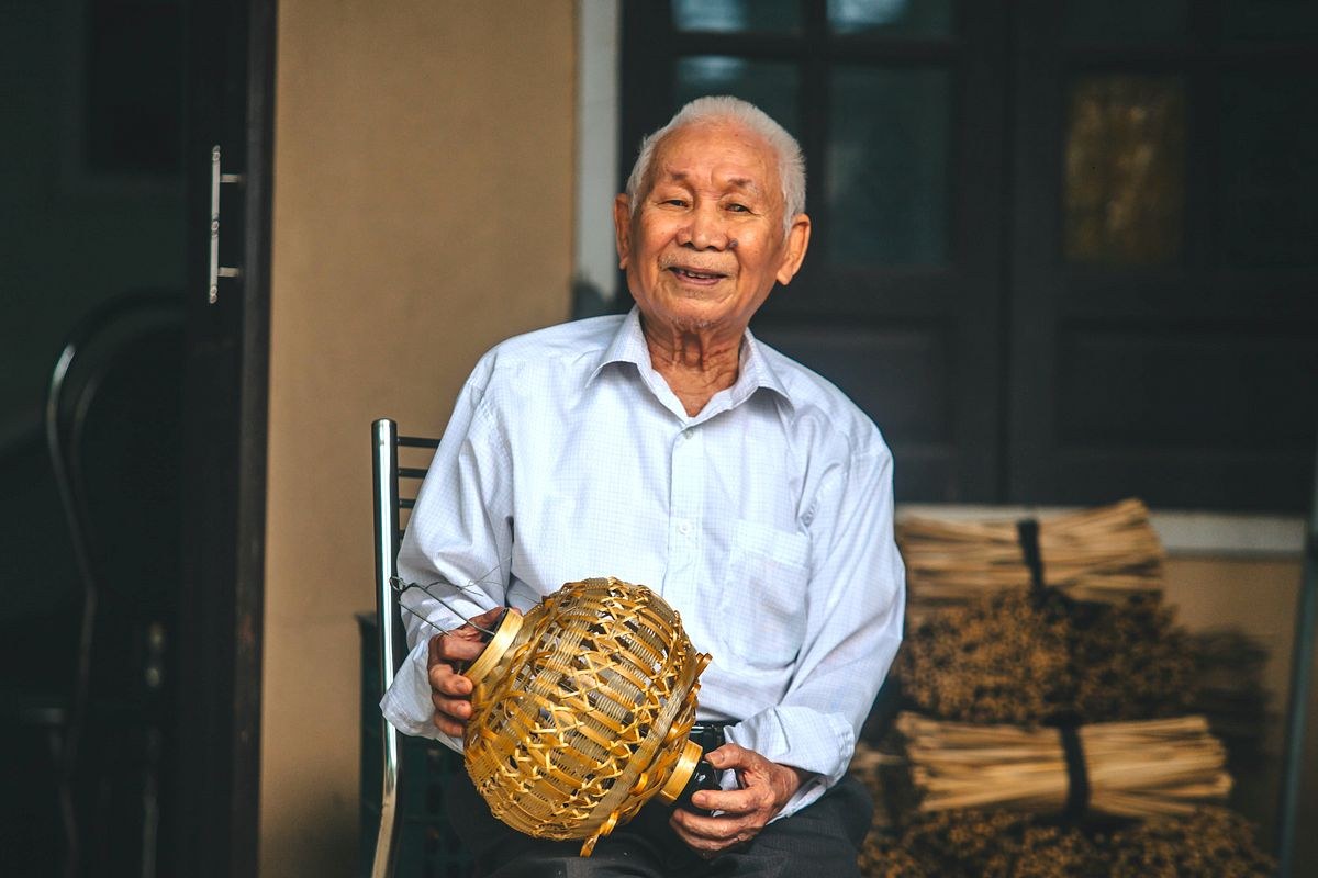 Portrait of artisan Huỳnh Văn Ba, his hair white, seated and smiling in a doorway, holding an unfinished round bamboo lantern frame in both hands, bundles of bamboo strips stacked behind him.