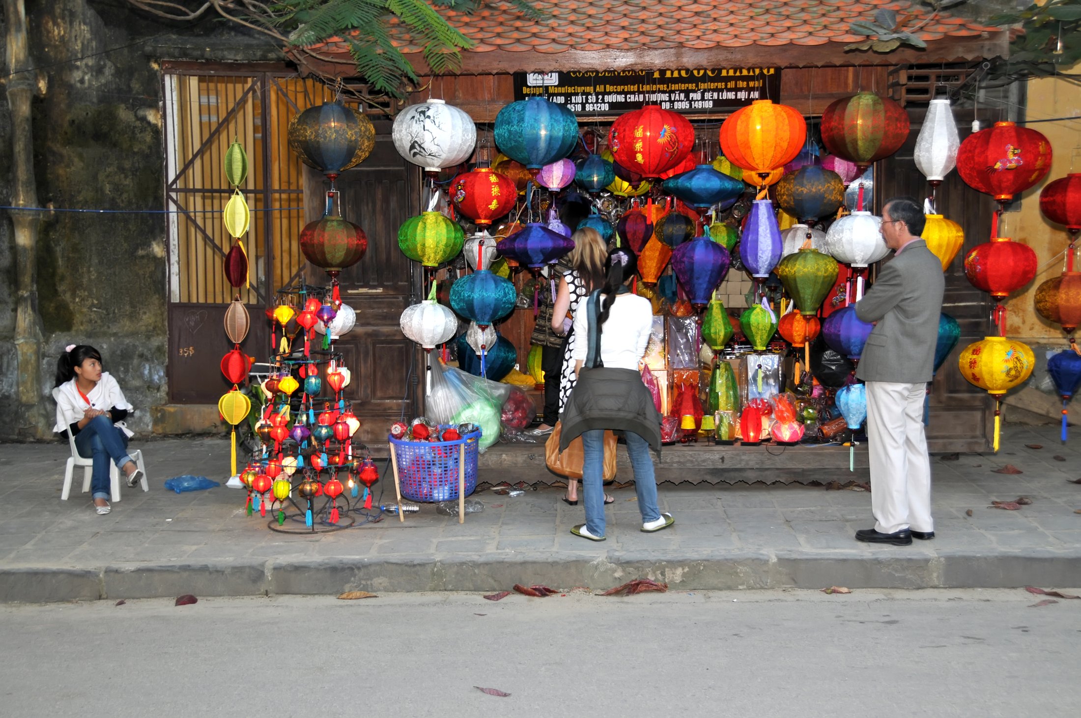 A lantern stall in Hội An at night, dozens of round and oval lanterns in red, orange, blue, purple and yellow hung across the whole front of a tiled wooden shopfront, two visitors and a vendor standing in front.