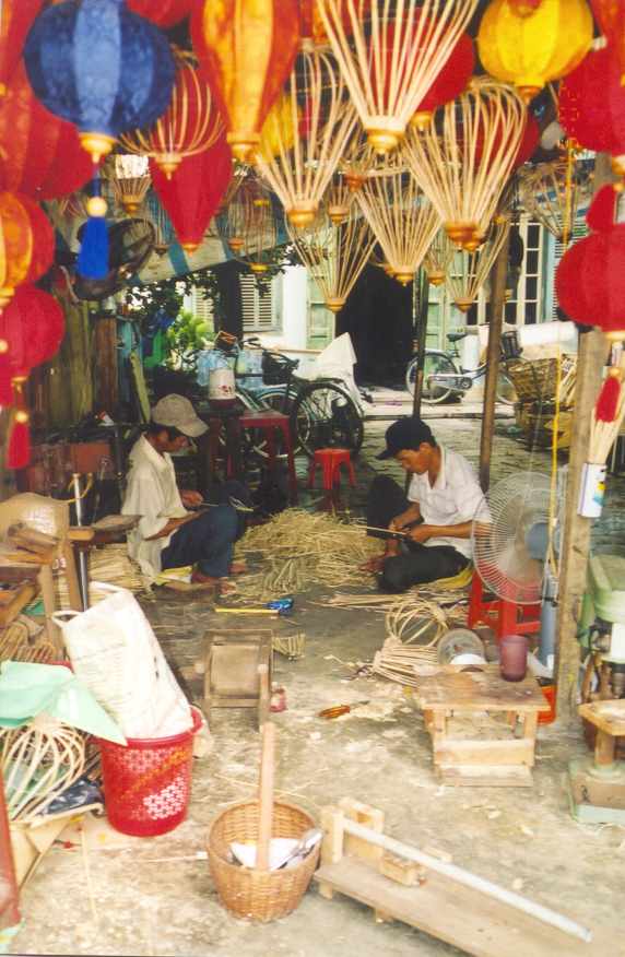 An open-air lantern workshop in Hội An: two men sit on the ground weaving bamboo frames, while dozens of finished, multi-coloured lanterns hang from the roof above them.