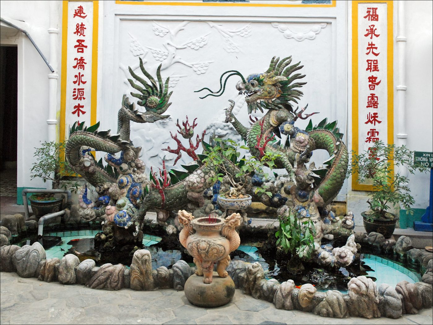 Con rồng khảm sành sứ và mảnh thủy tinh uốn khúc trên hồ nước nhỏ giữa chính điện và hậu điện, lư hương đá phía trước, câu đối chữ đỏ trên tường phía sau.