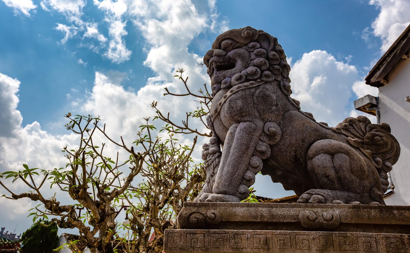 Sư tử đá phong hóa đứng trên bệ chạm ở lối vào hội quán, chụp từ dưới lên với nền trời sáng, bên cạnh là một cành sứ.
