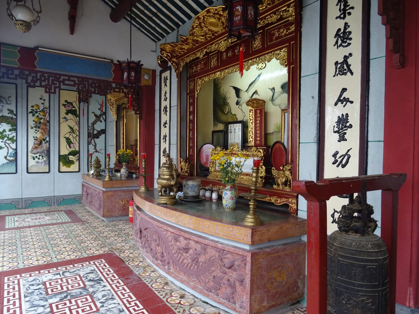 Một góc nhà Đông trong hội quán, nền lát gạch hoa văn, bàn thờ tiền hiền treo câu đối son thếp vàng, một quả chuông đồng cũ đặt trên giá đỏ phía trước.