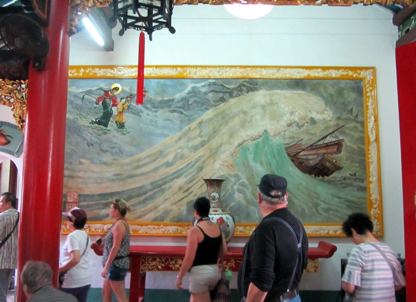 Tranh trên tường trong hội quán vẽ Thiên Hậu cùng một thị nữ bước trên sóng dữ, hướng về một chiếc thuyền gỗ đang chìm giữa phong ba, khách tham quan đứng xem phía trước.