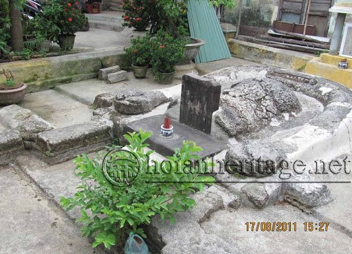 A small grave inside a house yard: a dark upright stele with a bundle of red incense sticks planted before it, potted flowers and stone steps around the grave, a corrugated roof behind. A timestamp in the corner reads 17/08/2011.