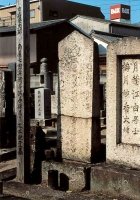 Two stone memorial markers standing side by side in the grounds of Raikō-ji, each with a small wooden nameplate above it, a vending machine behind them.
