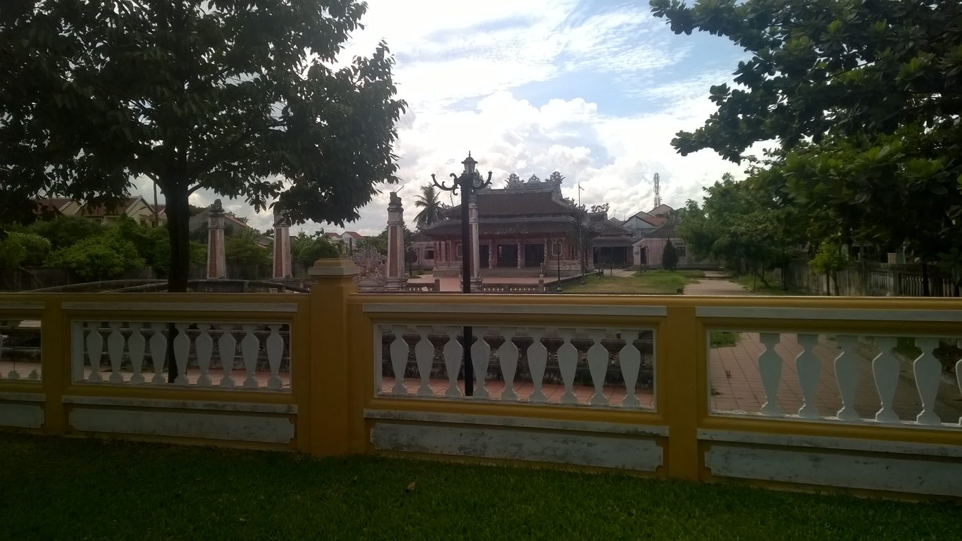 Một ngôi miếu mái ngói cong với trụ biểu hai bên sân, nhìn qua hàng rào con tiện sơn vàng và trắng, cây xanh hai bên, trời nhiều mây.