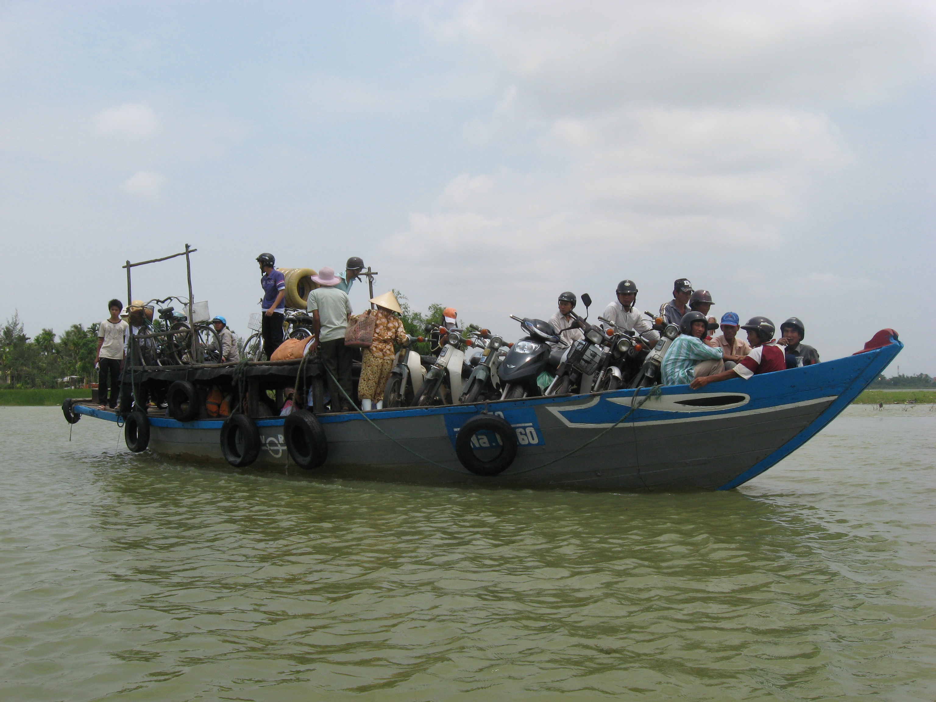 A crowded wooden ferry boat loaded with motorbikes, bicycles and passengers crossing the green water of the Thu Bồn river, rice fields and palm trees on the far bank.