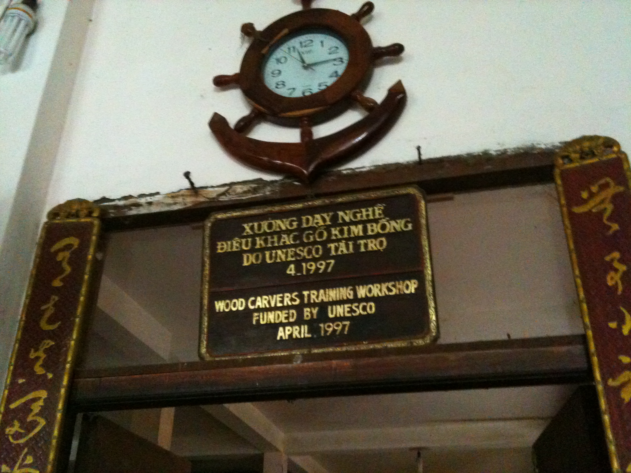 A bilingual wooden sign reading WOOD CARVERS TRAINING WORKSHOP FUNDED BY UNESCO APRIL 1997, mounted below a carved wooden ship's-wheel clock and anchor on a white wall.