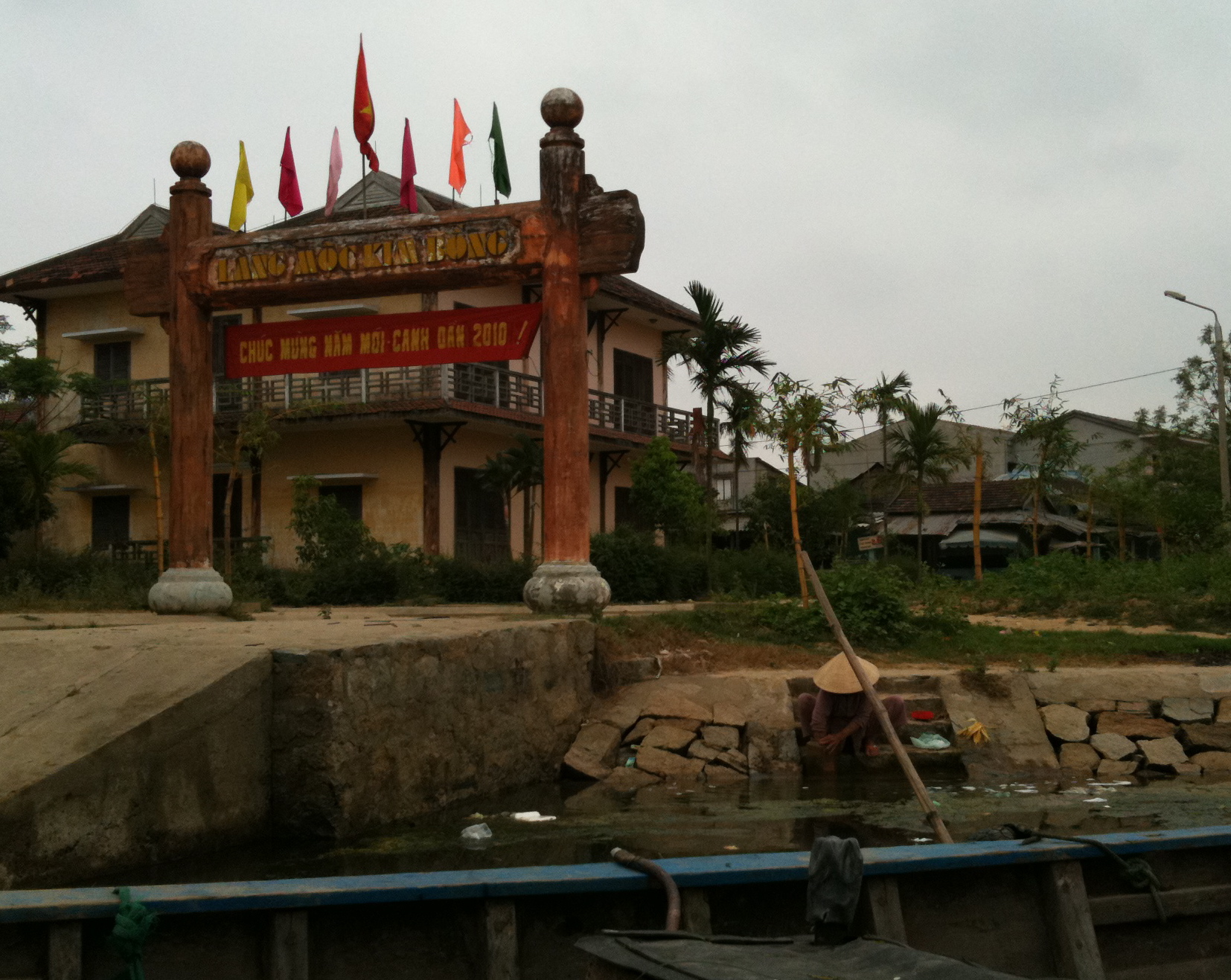 A timber gate reading LÀNG MỘC KIM BỒNG flying coloured flags and a Lunar New Year banner, a woman in a conical hat washing clothes at stone steps by the canal below, a boat's edge in the foreground.
