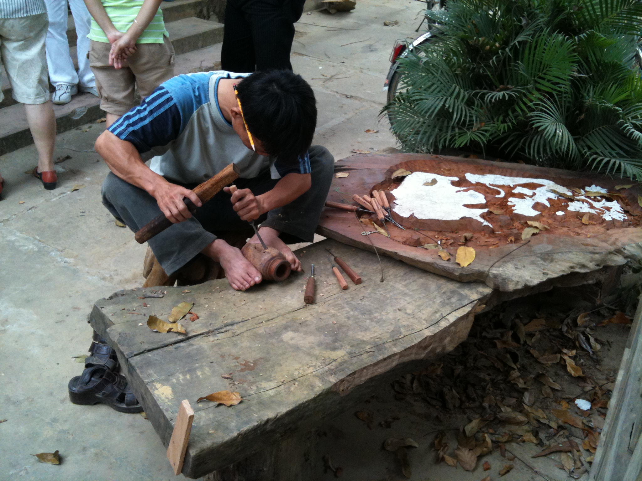 A young woodworker squats barefoot, gripping a piece of timber between his feet, striking a chisel with a wooden mallet, tourists watching from behind on a stone-paved yard scattered with wood shavings.