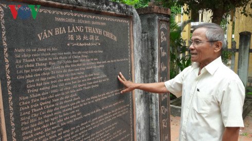 A stone stele carved with quốc ngữ and Chinese characters, titled "Văn bia làng Thanh Chiêm", an elderly man in a white shirt standing beside it, pointing at the inscription.