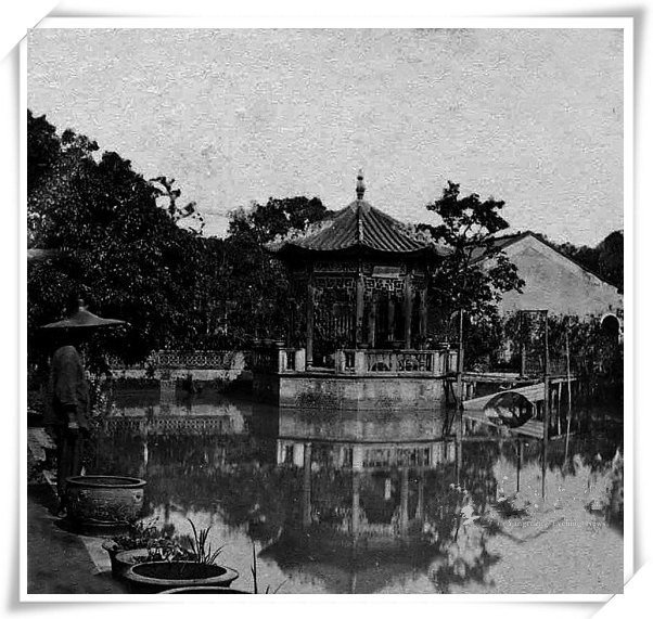 Ảnh cũ đen trắng: một đình mái ngói cong đứng giữa mặt ao lặng, phản chiếu xuống nước, cây cối rậm rạp bao quanh, một người đội nón đứng ở bờ ao phía trước.