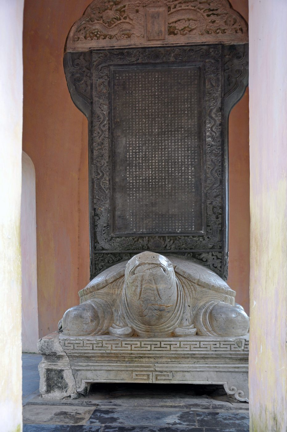 A large stone stele in a pink-walled pavilion: dragons carved across the head, the face densely cut with Chinese characters, coiled dragons along the border, the whole standing on the back of a white stone tortoise.