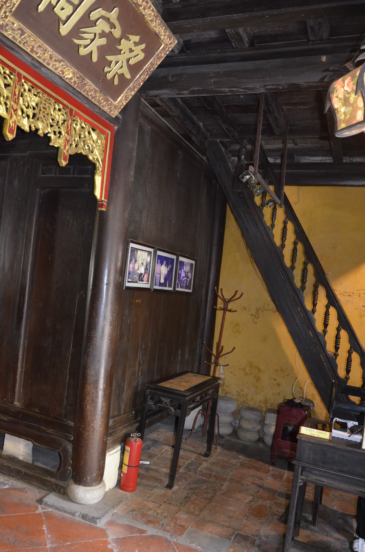 Cầu thang gỗ trong nhà cổ Tấn Ký đi lên cạnh các cột gỗ tròn đặt trên đá tảng, nền lát gạch phía dưới, một tấm hoành sơn son thếp vàng thấp thoáng phía trên bên trái.