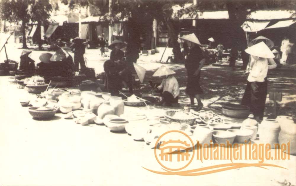An old sepia photograph of a street market with rows of earthenware pots, jars and bowls laid out on the ground, sellers and buyers in conical hats standing and crouching among the wares.