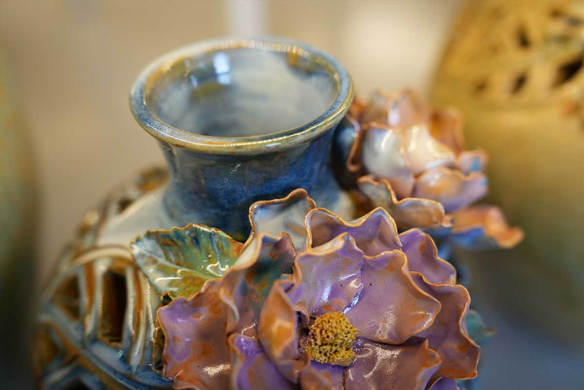 Close-up of a blue-glazed ceramic vase with raised, hand-formed purple and pink glazed flowers and green leaves decorating its neck.