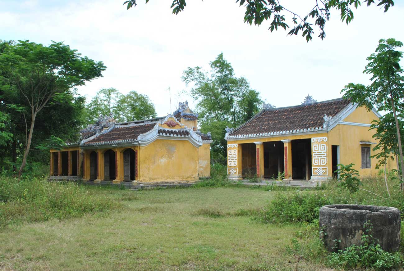 Two yellow-walled, tile-roofed shrine buildings of the Tổ Nghề temple complex in Nam Diêu, surrounded by grass and trees, an old stone-lined well visible at the lower right.