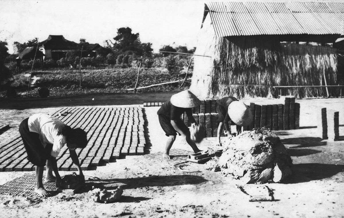 A black-and-white archival photograph of three people in conical hats making and drying roof bricks on a dirt yard beside a thatch-walled kiln shed, with rows of drying brick laid out on woven mats.