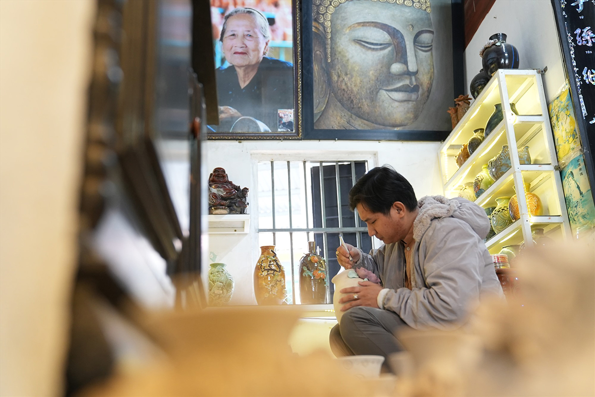 Nguyễn Viết Lâm bends over a glazed vase in his workshop painting decoration onto it, a photograph of an elderly woman and a painting of Buddha hanging on the wall behind him, shelves of colourful glazed pottery to the right.