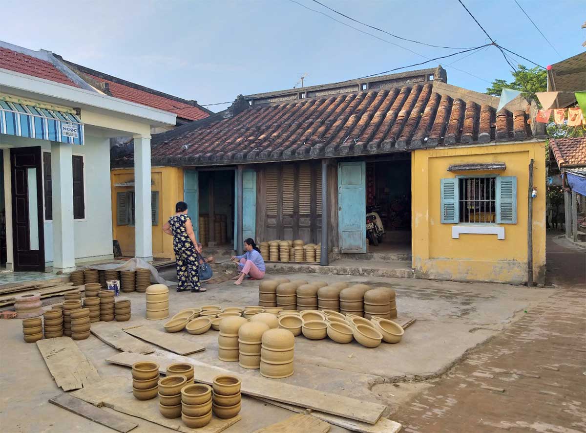 Unfired clay pots stacked on boards drying in front of a traditional yellow house with a tiled roof in Thanh Hà pottery village, two women working among the drying pots, more pots and wares visible through the open doorway.